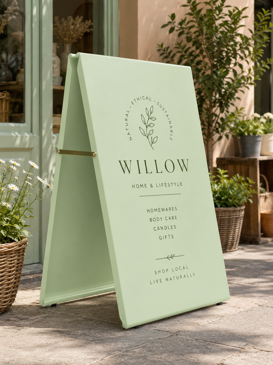 Pastel green RAL 6019 metal A-board pavement sign outside a modern lifestyle and homeware shop, featuring a minimalist botanical design with elegant typography. The soft mint green sign is displayed on a sunlit stone pavement surrounded by plants and natural décor, creating a premium boutique storefront aesthetic.
