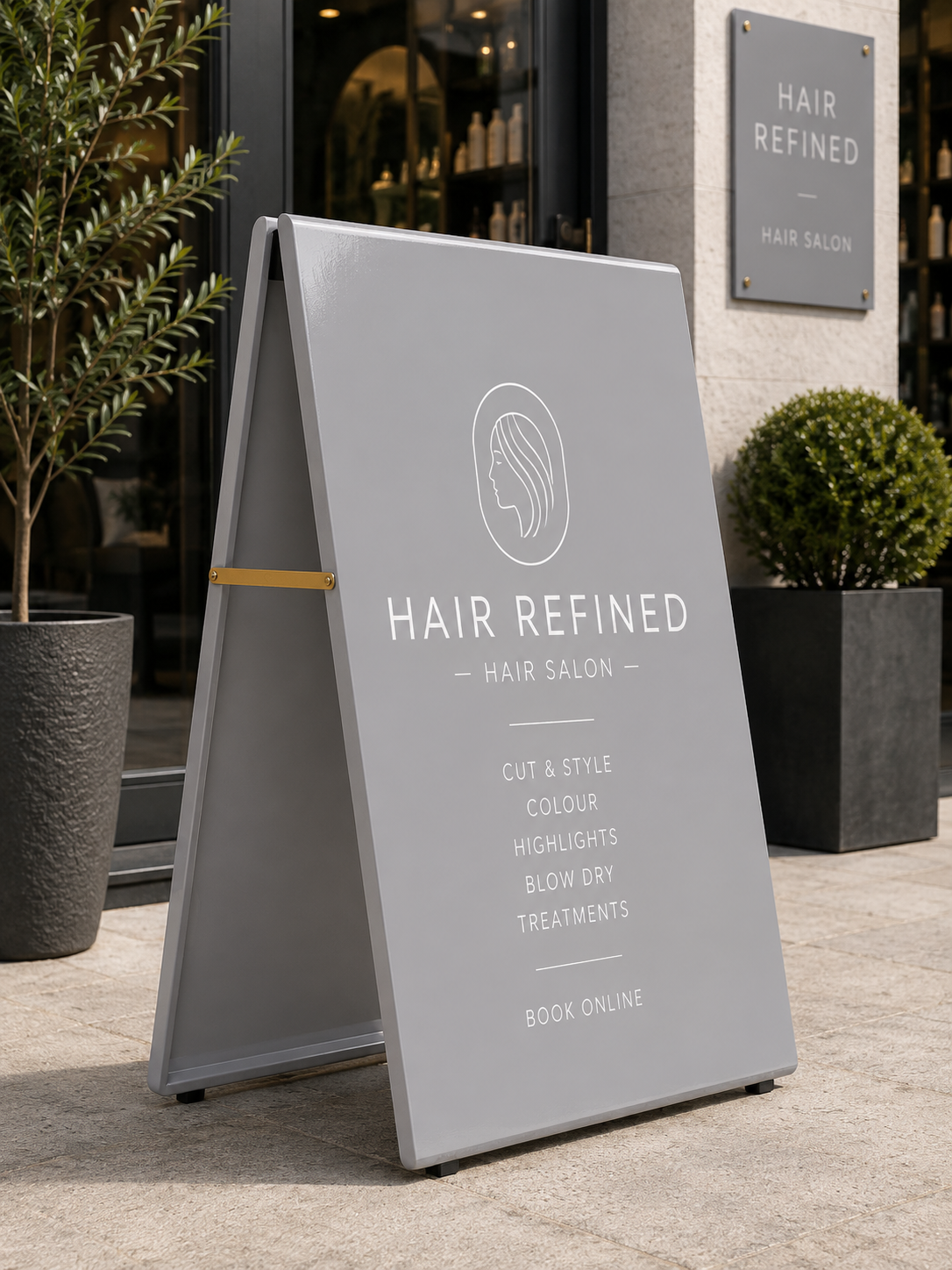 Platinum Grey RAL 7036 metal A-board pavement sign outside a contemporary hair salon, featuring minimalist white typography and elegant salon branding. The modern grey pavement sign is displayed on a light stone pavement with premium storefront styling, subtle greenery and luxury salon décor, creating a clean and sophisticated high-street retail aesthetic.