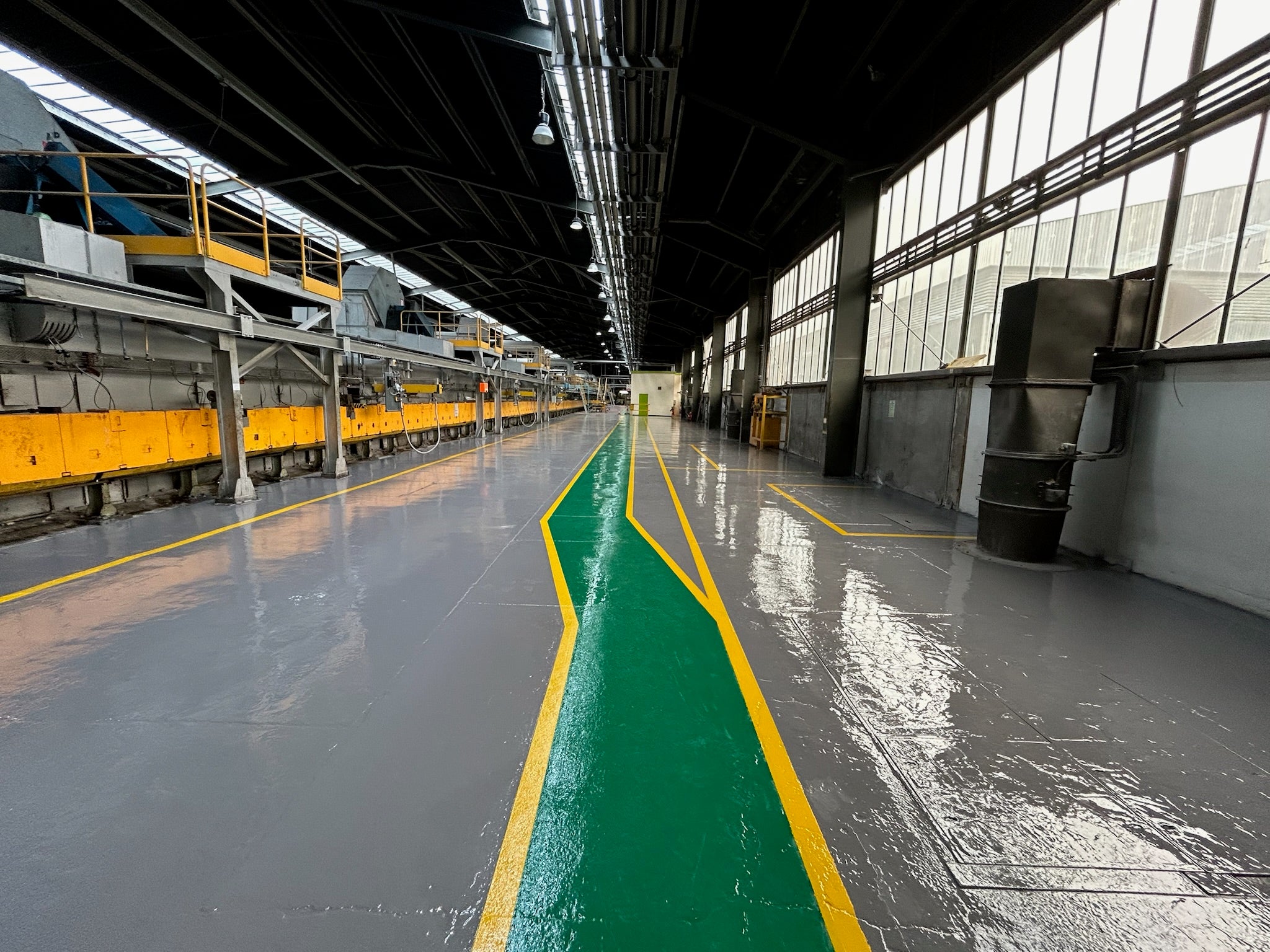 Industrial epoxy flooring in a factory with a glossy grey floor coating, green pedestrian walkway and yellow safety line markings.